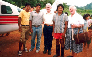 Three Waorani men, standing with Caucasian man and woman in front of an airplane on a jungle airstrip